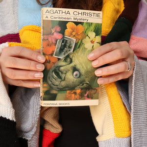alt="Cosy shot of hands holding a collectable copy of A Caribbean Mystery by Agatha Christie with artwork by Tom Adams. A knitted granny blanket can be seen draped over the shoulders of shop founder Bex Massey. Book and blanket are available from Bramble and Fox UK hygge shop."