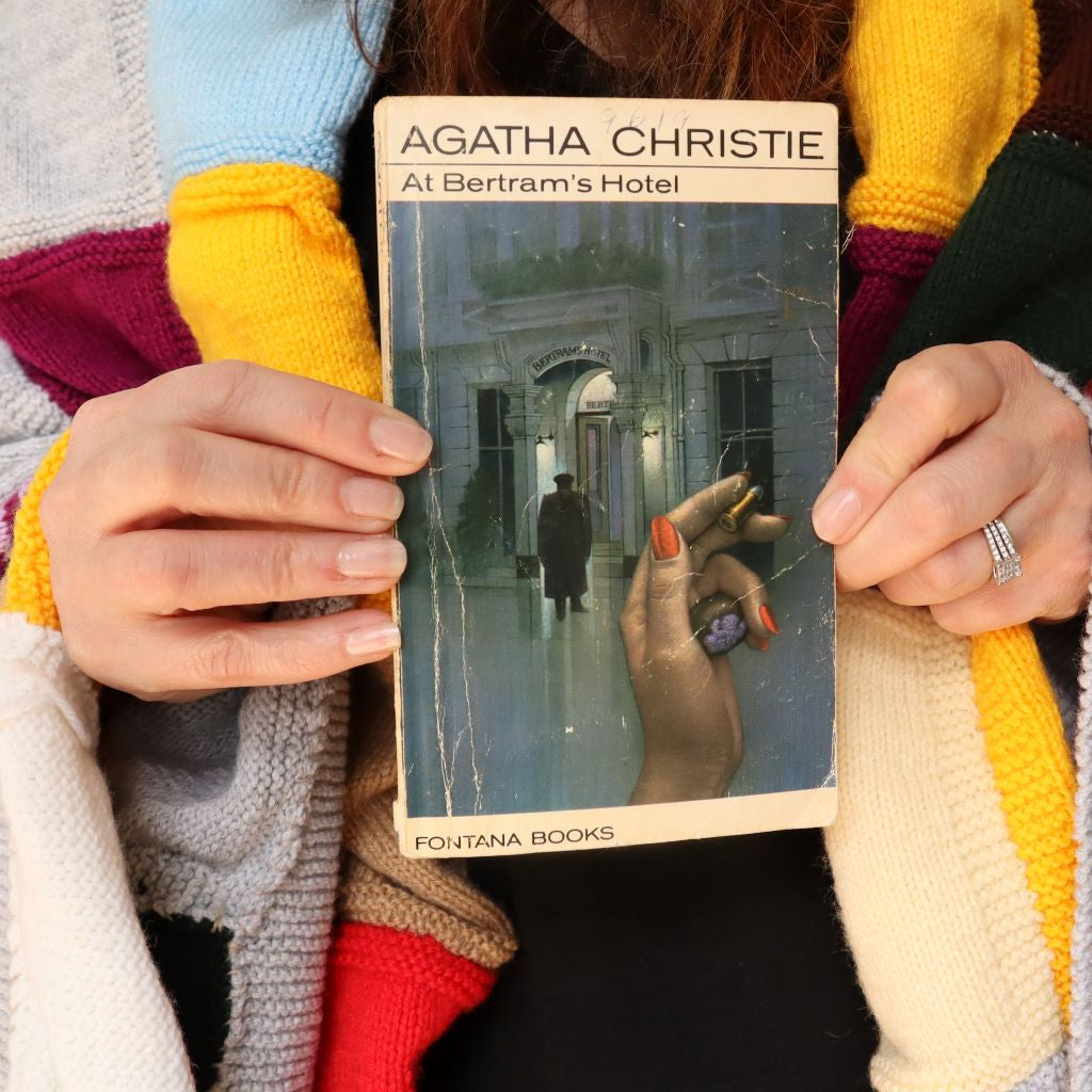 alt="Cosy shot of hands holding a collectable copy of At Bertam's Hotel by Agatha Christie with artwork by Tom Adams. A knitted granny blanket can be seen draped over the shoulders of shop founder Bex Massey. Book and blanket are available from Bramble and Fox UK hygge shop."