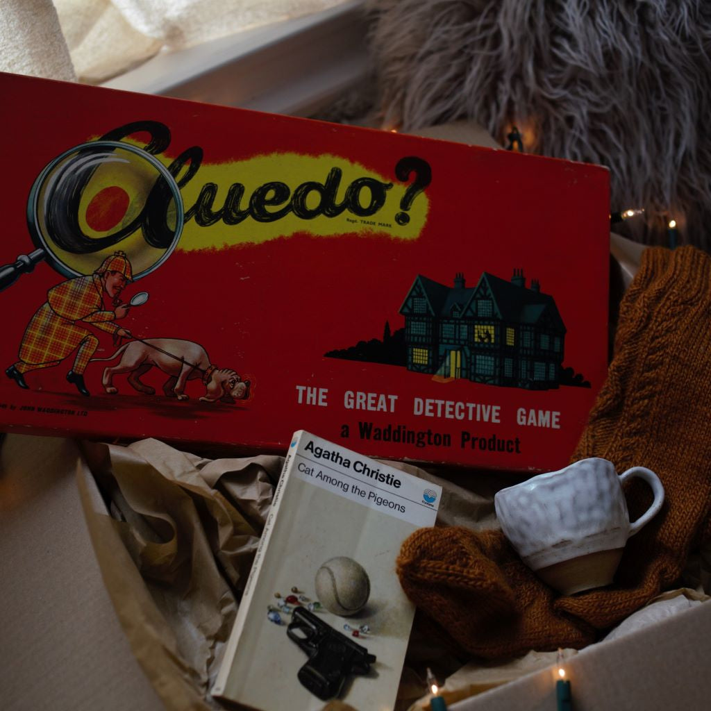 alt="cosy crime gift box featuring vintage Cluedo, vintage Agatha Christie novel, cream handmade mug, handknitted woollen socks. Available from Bramble and Fox UK Hygge Shop."