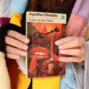 alt="Cosy shot of hands holding a collectable copy of Taken at the Flood by Agatha Christie with artwork by Tom Adams. A knitted granny blanket can be seen draped over the shoulders of shop founder Bex Massey. Book and blanket are available from Bramble and Fox UK hygge shop."