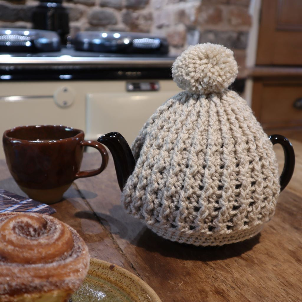 alt="cream crochet knit tea cosy with pom pom, on a brown betty tea pot. A brown handmade cup and studio pottery plate with cinnamon bun are in foreground. A cream AGA can be seen in background. Hygge homeware available from Bramble and Fox UK hygge shop"