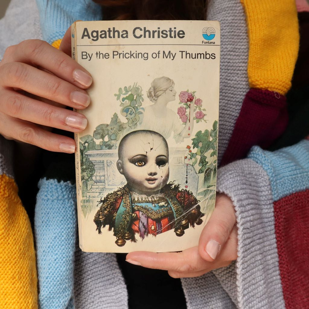 alt="Cosy shot of hands holding a collectable copy of By The Pricking of My Thumbs by Agatha Christie with artwork by Tom Adams. A knitted granny blanket can be seen draped over the shoulders of shop founder Bex Massey. Book and blanket are available from Bramble and Fox UK hygge shop."