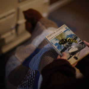 alt="Cosy POV shot of hands holding a collectable copy of Death in the Clouds by Agatha Christie with artwork by Tom Adams. Outstretched legs covered by a knitted granny blanket can be seen. Book and blanket are available from Bramble and Fox UK hygge shop."