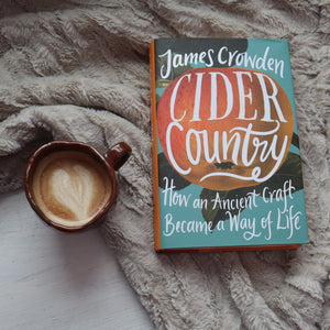 alt="cider country book by james crowden resting on a scandi faux fur blanket. A brown handmade mug with latte art sits next to it. Available from Bramble and Fox uk hygge homeware shop"