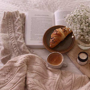 alt="hygge breakfast flatlay featuring ochre and flax candle, cream handmade pottery mug, cream hand knitted socks, studio pottery plate and Nigel slater A cook's book. All available from Bramble and Fox UK hygge homewares."