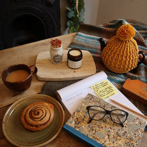 alt="hygge tablescape featuring ochre and flax candle, numol jar, brown handmade pottery mug, graphis books journal and tartan Tweedmill blanket. All available from Bramble and Fox UK hygge shop"