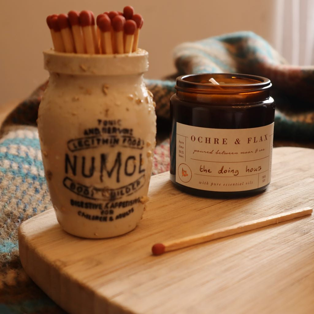 alt="the doing hours aromatherapy candle by ochre and flax sitting on a wooden board with a numol jar filled with matches. All available from Bramble and Fox UK hygge homewares"
