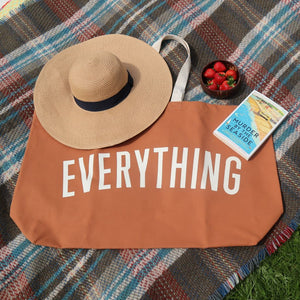 alt="tan canvas beach bag featuring the word EVERYTHING in white uppercase letters. Flatlay includes a sun hat, crime novels, a bowl of strawberries. Bag made by Alphabet Bags. Available from Bramble and Fox UK hygge gifts and homewares"