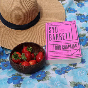 alt="flatlay featuring book Syd Barrett A Very Irregular Head by Rob Chapman, sun hat, coconut shell bowl of strawberries and blue flower power psychadelic fabric. Book, bowl and fabric available from Bramble and Fox UK cottagecore home and gift shop"