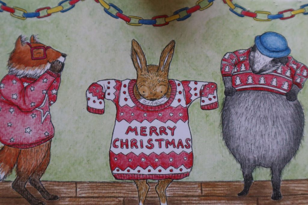 alt="close up of The Wrong Christmas Jumpers christmas card by Kerry Dilks. Featuring badger squeezing into a too tight christmas jumper and rabbit in a huge christmas jumper with fox at the side chuckling. Cosy illustration in the style of Raymond Briggs and Janet Ahlberg. Available from Bramble and Fox UK hygge homewares and gifts"