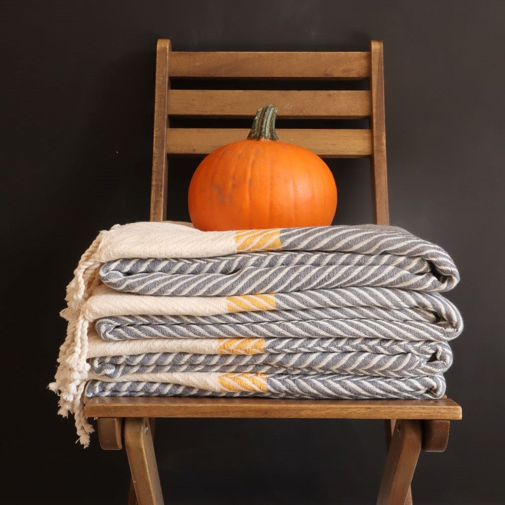 alt="a stack of mustard grey herringbone throws on a wooden chair, crowned by a pumpkin. Perfect for Autumn. Available from Bramble and Fox UK hygge home decor"