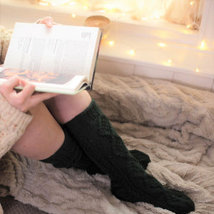 alt="woman's legs in green knitted woollen socks. She's reading a book with a mug, blanket and fairylights. Socks available from Bramble and Fox UK hygge homewares"