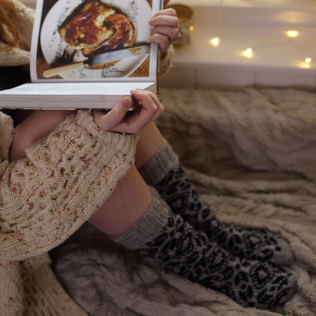 alt="woman in chunky knit cardigan reading book wearing knitted grey woollen socks with fair isle design and fairy lights in background. Socks available from Bramble and Fox UK hygge homewares and gifts."