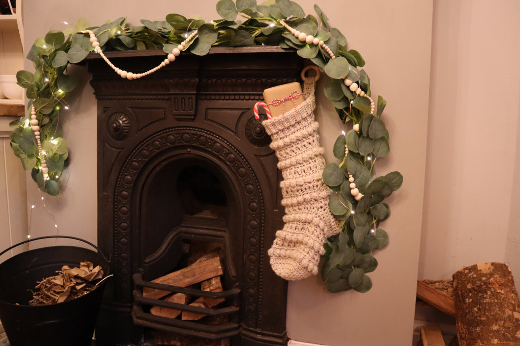 alt="cast iron victorian fireplace filled with logs and decorated with cream hand knitted christmas stocking and eucalyptus garland. Stocking available from Bramble and Fox UK hygge gifts and homeware."