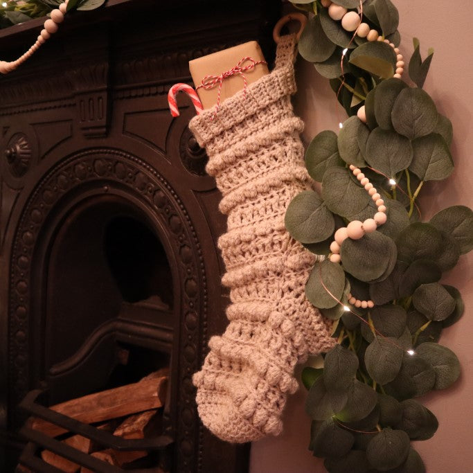 alt="cream bobble stitch, popcorn knit christmas stocking hanging on a victorian fireplace with garland. Available from Bramble and Fox UK hygge gifts and homeware"
