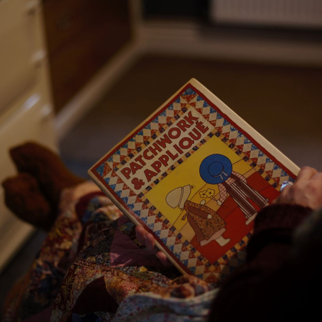 alt="golden hands patchwork and applique book by pamela tubby. woman's hands holding book so cover is visible to camera. Book is resting on outstretched legs covered with a patchwork quilt. Book and patchwork blanket available from Bramble and Fox UK hygge interiors shop"