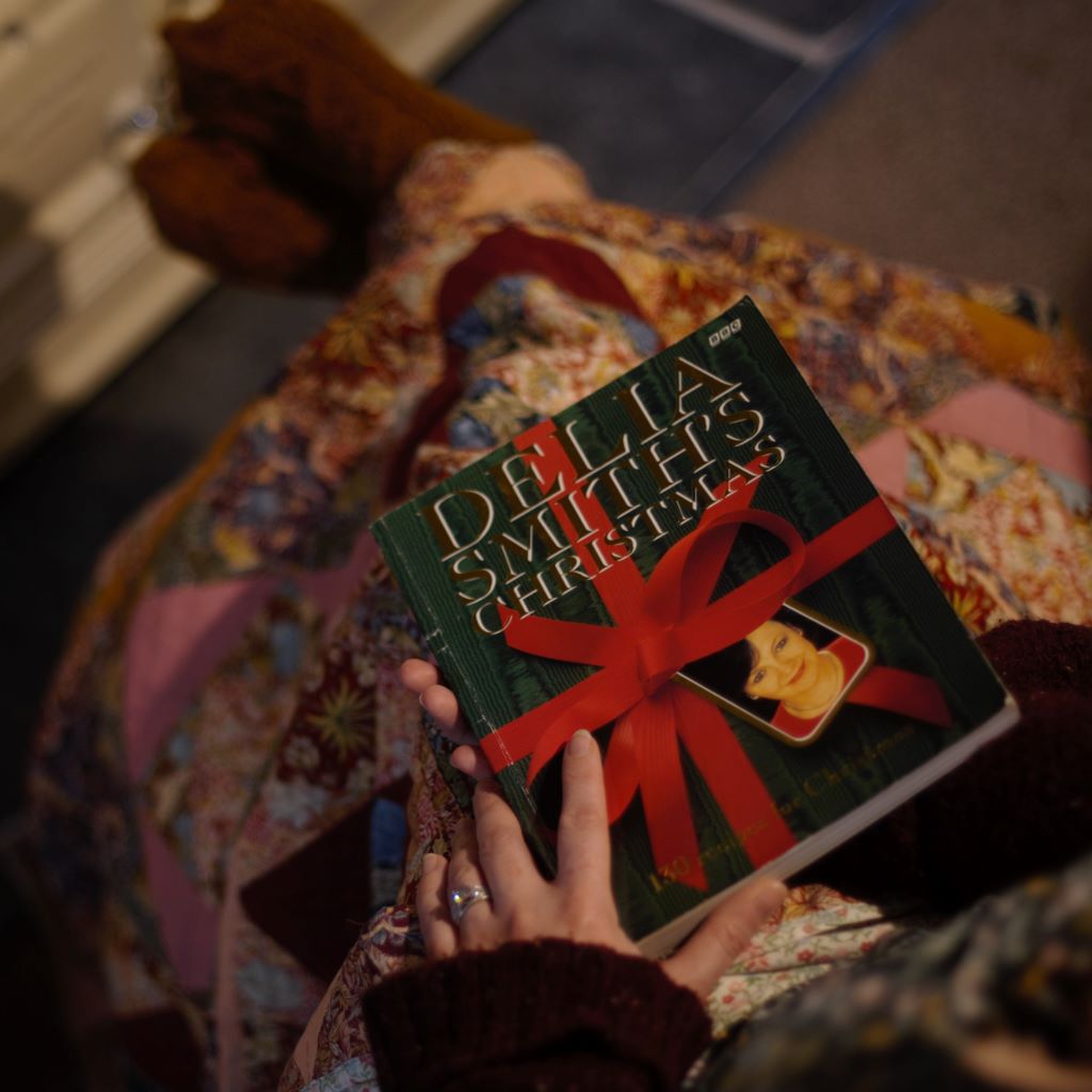 alt="POV shot of woman's legs covered with patchwork blanket. Hands holding a copy of Delia Smith's Christmas. Book available from Bramble and Fox Uk hygge homeware and gifts"
