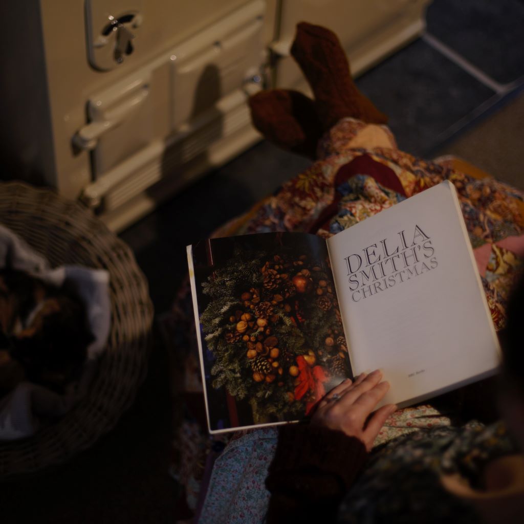 alt="woman snuggled up next to aga with patchwork blanket, reading Delia Smith's Christmas. Book available from Bramble & Fox UK hygge homeware and gifts"