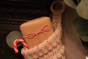 alt=brown paper parcel wrapped with baker's twine and a candy cane peeking out of a cream cable knit crhistmas stocking hanging from a victorian fireplace. Available from Bramble and Fox UK hygge gifts and homeware"