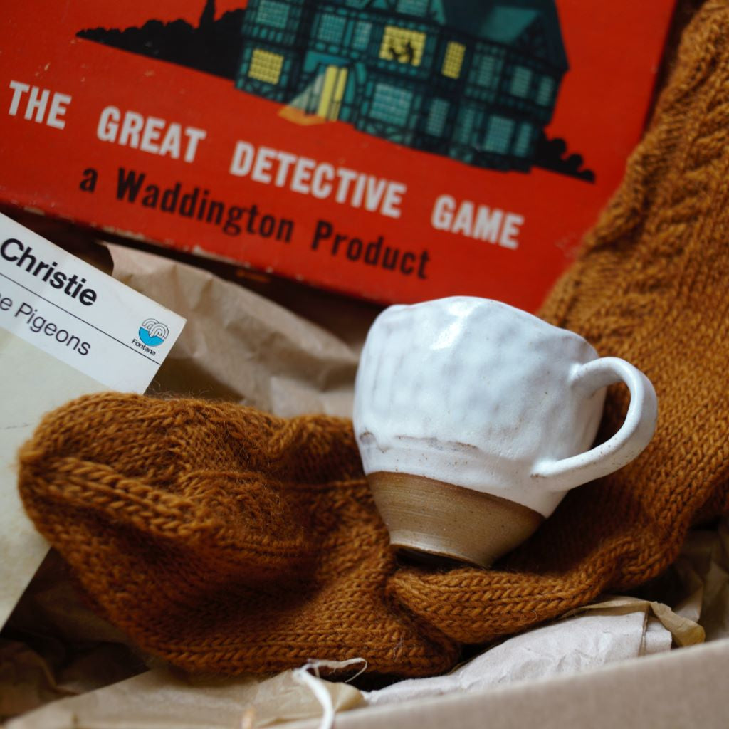 alt="tactile handmade cream glazed pinch pot mug resting on top of gingerbread coloured hand knitted socks. Vintage Cluedo in background. Cosy Crime box available from Bramble and Fox UK hygge homewares"