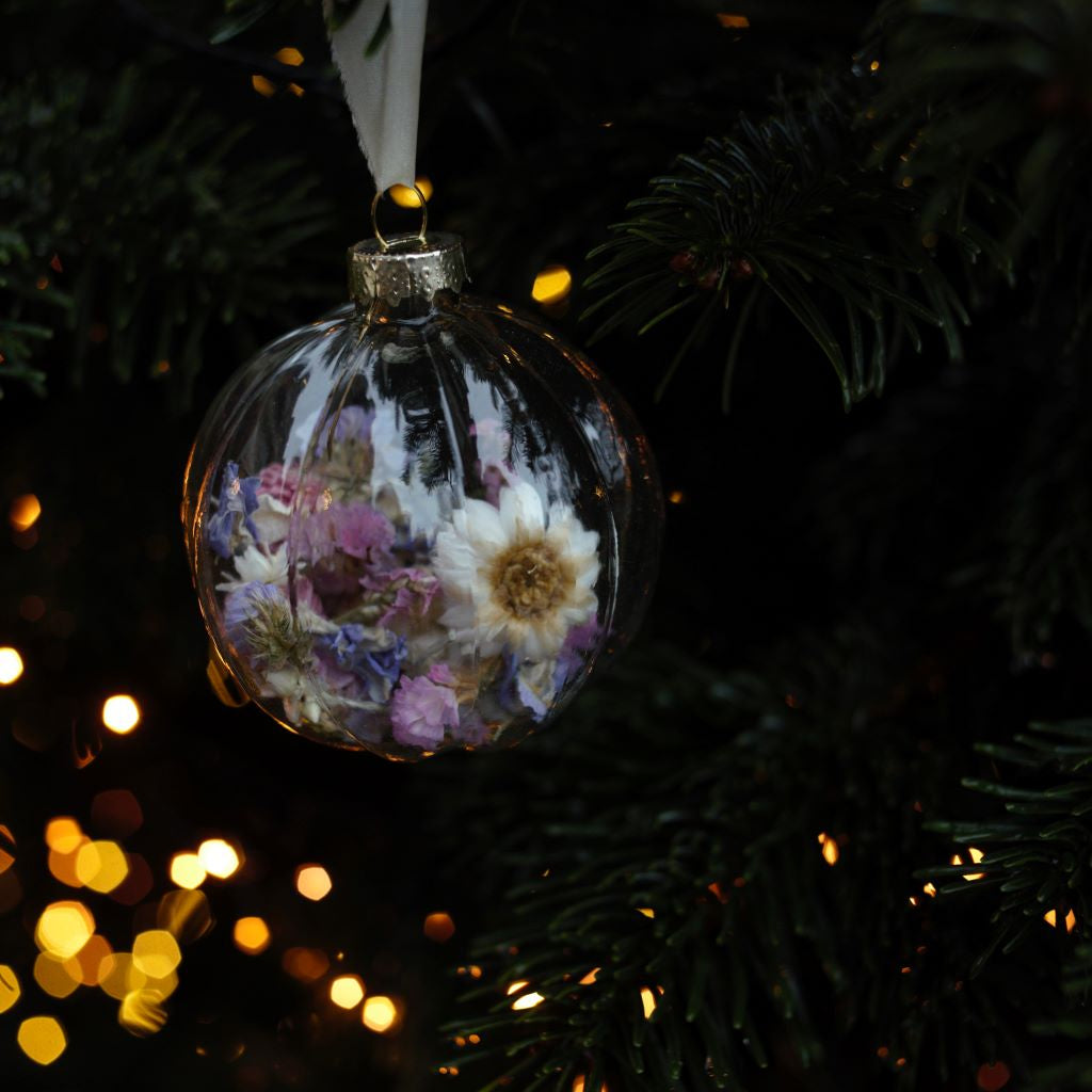 alt="glass christmas bauble filled with dried flowers hanging from Christmas tree branch with fairy lights in backround. Available from Bramble and Fox UK hygge homeware shop"