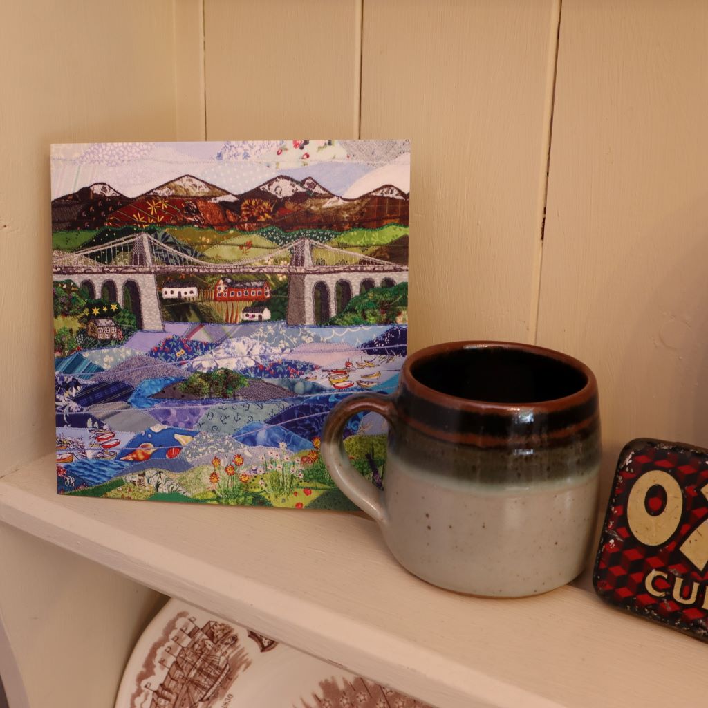 alt="menai bridge patchwork style card on an old dresser with handmade mug. Available from Bramble and Fox UK hygge homeware shop"