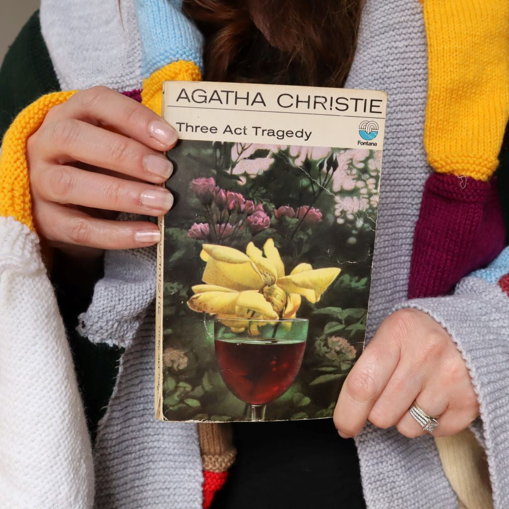 alt="Cosy shot of hands holding a collectable copy of Three Act Tragedy by Agatha Christie with artwork by Tom Adams. A knitted granny blanket can be seen draped over the shoulders of shop founder Bex Massey. Book and blanket are available from Bramble and Fox UK hygge shop."