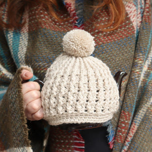 alt="cream crochet knit tea cosy with pom pom keeps a Cauldon Brown Betty teapot snug and warm.  A woman draped in a Tweedmill tartan blanket is holding the teapot. Teapot, tea cosy and blanket are available from Bramble and Fox UK hygge homewares and gifts shop"